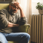 A weary man, a caregiver, sits in a chair with his head in his hands, reflecting his fatigue and stress.
